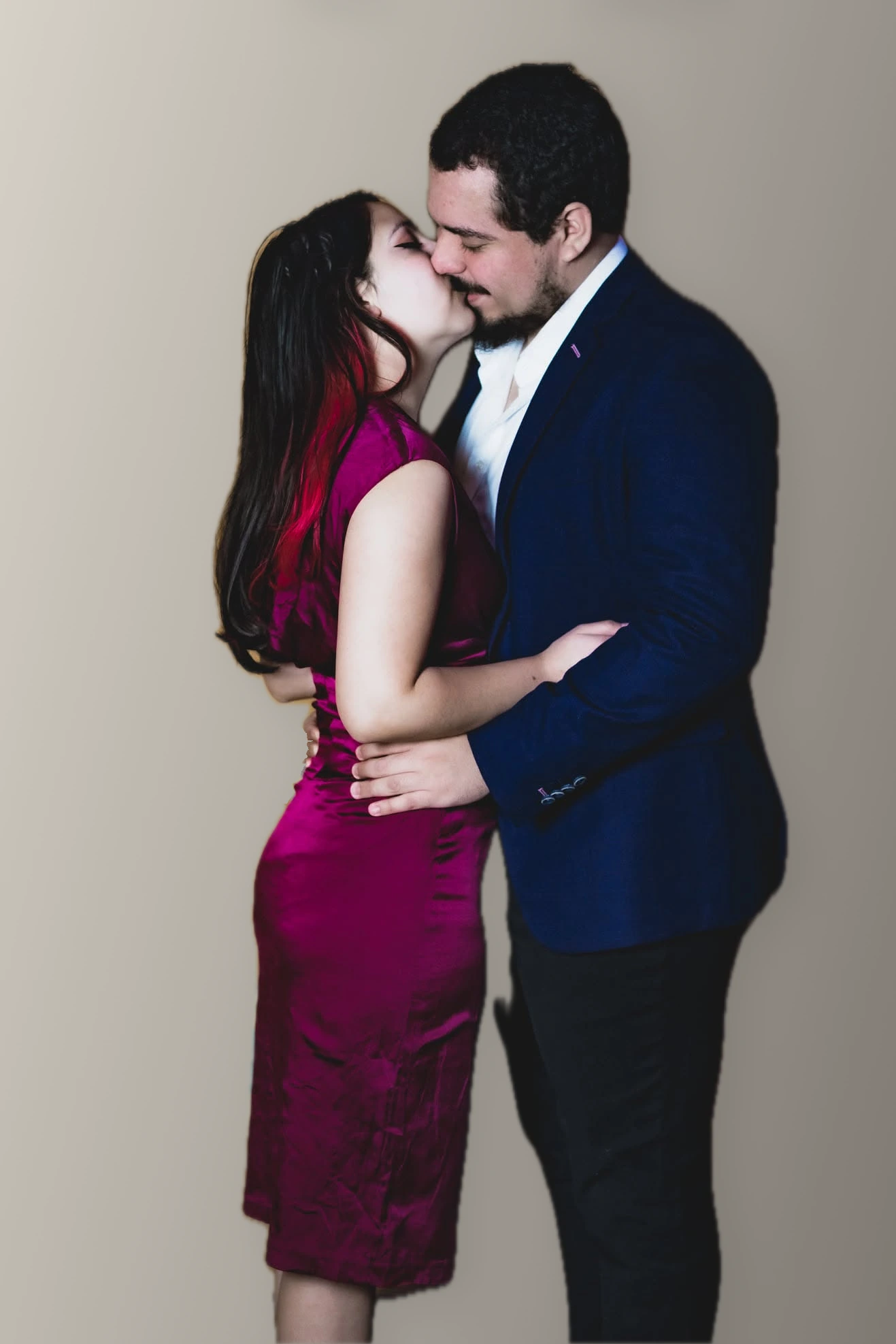Josefa y Gabriel besándose, con vestido magenta y traje azul, ante un fondo neutro cálido.